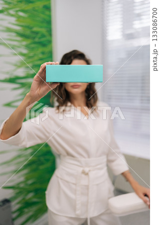Young woman ophthalmologist in white coat holding a light-blue product box over her eyes in a modern clinic, symbolizing eye care, vision solution advertising and medical wellness 133086700