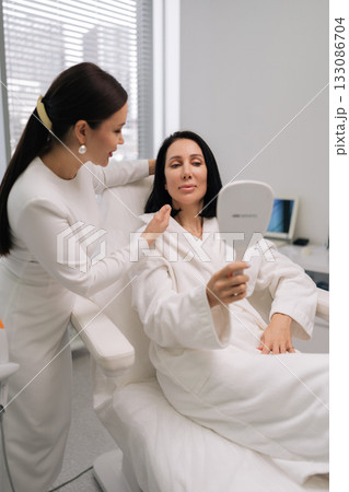 Vertical shot of beautician providing personalized consultation to female client in specialized treatment chair, reviewing her skin appearance in mirror inside contemporary beauty salon. Vertical shot of beautician providing personalized consultation to female client in specialized treatment chair, reviewing her skin appearance in mirror inside contemporary beauty salon. 133086704