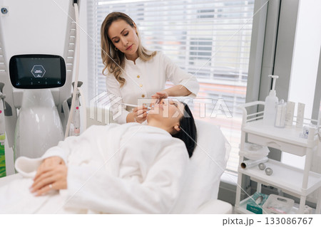 Professional beautician preparing female client for facial cosmetic procedure, using skin marking pencil and stencil to outline treatment areas for enhanced beauty. Concept of facial anti-aging. Professional beautician preparing female client for facial cosmetic procedure, using skin marking pencil and stencil to outline treatment areas for enhanced beauty. Concept of facial anti-aging. 133086767