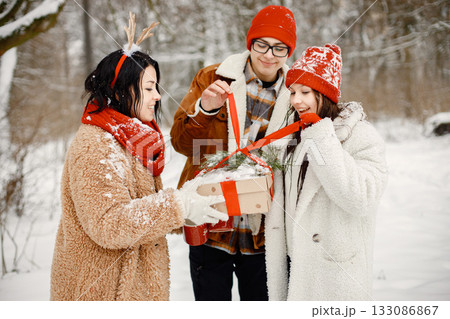 Joyful teens and their beautiful mother spending time together in the winter park. Family holding a gift box. Family wearing warm jackets and knitted hats. 133086867