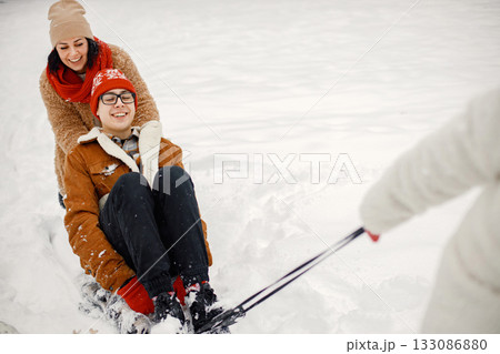 Joyful boy and his beautiful mother spending time together in the winter park. Woman wearing warm jacket and red sweater. Family having fun riding a sled. Joyful boy and his beautiful mother spending time together in the winter park. Woman wearing warm jacket and red sweater. Family having fun riding a sled. 133086880
