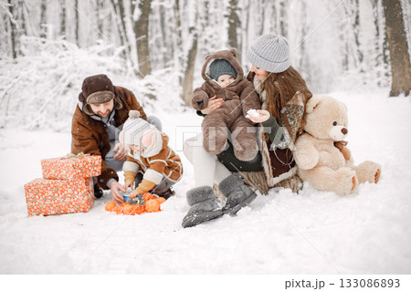Parents with their children having fun in forest on snow day. Happy toddler daughter and baby son wearing warm clothes. Gift boxes standing next to them. 133086893