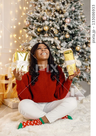 Black woman sitting near Christmas tree at home. Woman opening a gift in golden paper. Brunette girl wearing red sweater and white trousers. 133086933