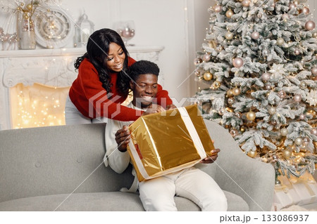 Young black couple with gifts in Christmas eve. Man sitting on a coach at home. Brunette woman standing behind and presented a gift for her boyfriend. Young black couple with gifts in Christmas eve. Man sitting on a coach at home. Brunette woman standing behind and presented a gift for her boyfriend. 133086937