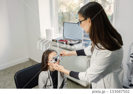 Little girl checking up her sight at ophthalmology center. Female optometrist doing sight testing. Brunette girl wearing grey jacket and black shirt. 133087502