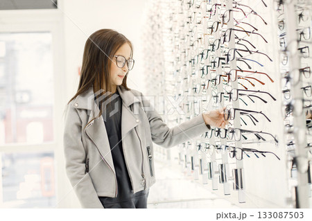 Little girl choosing eyeglasses in optica shop. Female customer taking eyeglasses from shelves in optica. Girl wants to try eyeglasses on. 133087503