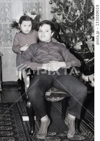 A vintage portrait of a sister and brother near a New Year's tree. A girl with bows hugs her brother. Retro 1973 A vintage portrait of a sister and brother near a New Year's tree. A girl with bows hugs her brother. Retro 1973 133089019