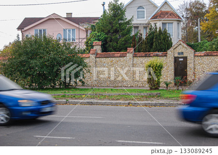 Residential street scene with stone wall and passing vehicles. Residential street with a tall stone wall featuring red-tiled caps. The street is occupied by passing blurry blue cars. Residential street scene with stone wall and passing vehicles. Residential street with a tall stone wall featuring red-tiled caps. The street is occupied by passing blurry blue cars. 133089245