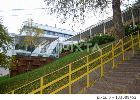 Modern building on a grassy hill with yellow railing and steps. Modern building with white and glass facade, featuring angular design elements.  133089452