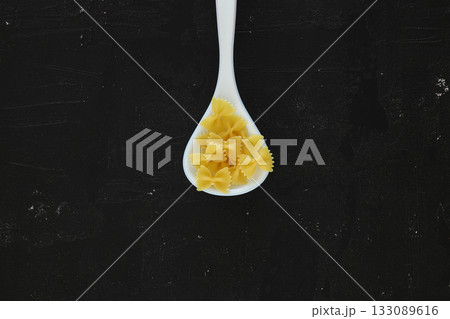 Raw pasta farfalle on wooden spoon on black background. Top view of Italian cuisine ingredient. 133089616