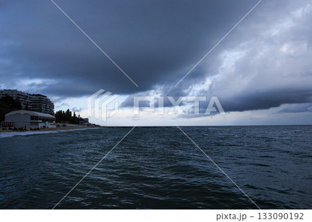 View of the coastal slopes of Odessa with a dark bay and cloudy sky. Dark, cloudy sky looms over a seaside town along the coast. The sea is deep and choppy, stretching towards the horizon.  133090192