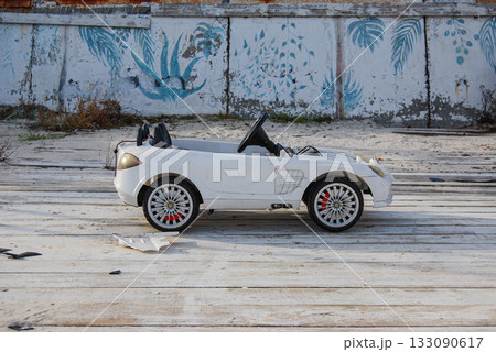 An abandoned white toy car against a concrete wall with leaf patterns. A small white toy convertible car sits on a worn wooden deck on a sandy beach. 133090617