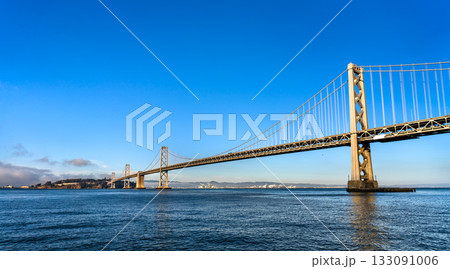 A wide, low-angle view of the San Francisco-Oakland Bay Bridge from the water. The structure spans the bay under a clear, vibrant blue sky. This image represents travel, engineering, and Bay Area 133091006