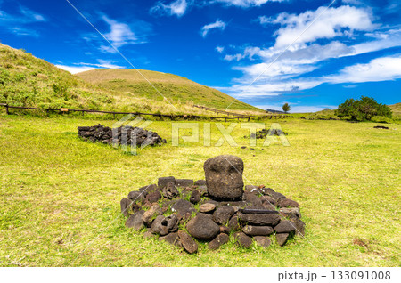 View of an archaeological site at Anakena Beach, Easter Island. A stone head is surrounded by rocks on a grassy hill under a blue sky 133091008