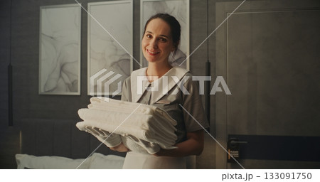 Smiling Woman in Neat Professional Uniform Holds Folded Stack of Fresh White Towels in Hotel Room Smiling Woman in Neat Professional Uniform Holds Folded Stack of Fresh White Towels in Hotel Room 133091750