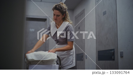 Female Hotel Housekeeper in Neat Uniform Stands Beside Cleaning Trolley, Carefully Folding Towels 133091764
