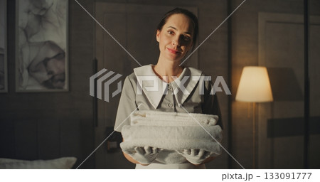 Hotel Maid in Classic Uniform With White Gloves Holds Clean Soft Towels, Standing in Hotel Bedroom Hotel Maid in Classic Uniform With White Gloves Holds Clean Soft Towels, Standing in Hotel Bedroom 133091777