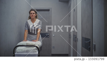 Female Maid in Professional Uniform Moves Cleaning Trolley Down Hallway, Carrying Neatly Towels Female Maid in Professional Uniform Moves Cleaning Trolley Down Hallway, Carrying Neatly Towels 133091799