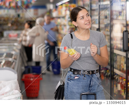 Young woman chooses sweet drink in a glass bottle in the grocery section of supermarket 133094015