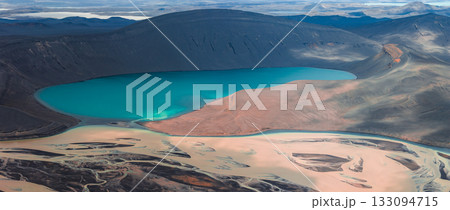 Turquoise crater lake surrounded by dark volcanic terrain, sandy delta, braided rivers, rugged hills, and distant mountains in Iceland. 133094715