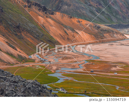 Vibrant orange and brown rhyolite hills with green mossy plains, winding streams, and a road with parked cars in Landmannalaugar, Iceland. Vibrant orange and brown rhyolite hills with green mossy plains, winding streams, and a road with parked cars in Landmannalaugar, Iceland. 133094719