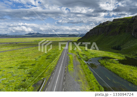 A paved road extends through a green landscape in Iceland, bordered by a stream and grassy hillside, with mountains and clouds in the distance. A paved road extends through a green landscape in Iceland, bordered by a stream and grassy hillside, with mountains and clouds in the distance. 133094724