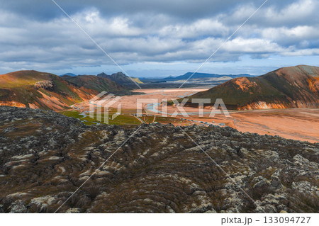 Aerial view of Iceland's Landmannalaugar region with vibrant rhyolite mountains, dark lava fields, a winding river, and a cloudy sky in the highlands. 133094727