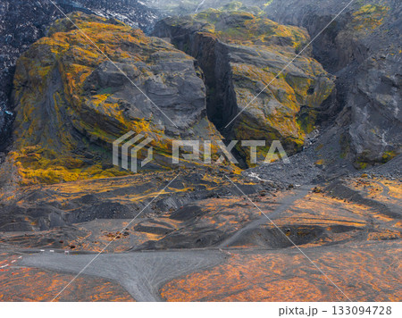 Steep volcanic cliffs with yellow and green moss, black soil, and reddish vegetation. A dirt road winds through the dramatic Icelandic landscape. 133094728