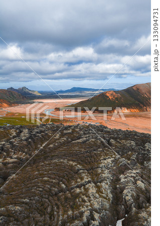 Aerial view of Iceland's Landmannalaugar region with vibrant rhyolite hills, dark lava fields, a winding river, and an overcast sky. 133094731
