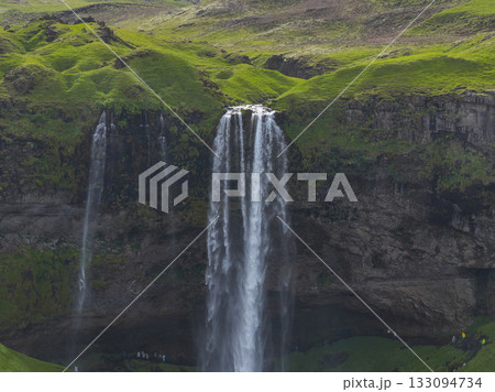 Seljalandsfoss waterfall in Iceland cascades from a tall cliff, surrounded by green grass and mossy rocks, with a visible pathway behind the water. Seljalandsfoss waterfall in Iceland cascades from a tall cliff, surrounded by green grass and mossy rocks, with a visible pathway behind the water. 133094734