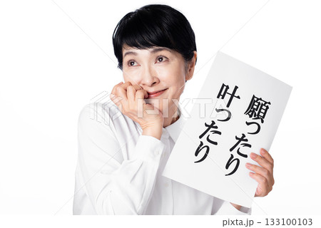 願ったり叶ったりと書かれた紙を持ち意味深げに笑う白いシャツを着たシニア女性 133100103