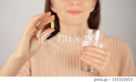 Macro close-up of an unrecognizable smiling woman holding vitamin capsule and glass of water. Advertising concept for vitamins and nutritional supplements, health care. Macro close-up of an unrecognizable smiling woman holding vitamin capsule and glass of water. Advertising concept for vitamins and nutritional supplements, health care. 133103053