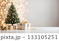 Elegant Christmas tree with golden ornaments beside wrapped gift boxes on pristine white surface with warm bokeh lighting background 133105251