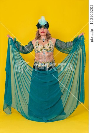 Caucasian woman fashion model with outstretched arms attired in green eastern dance stage outfit pareo, turban, bra top and maxi skirt with slit and she is not wearing footwear on yellow background 133106203