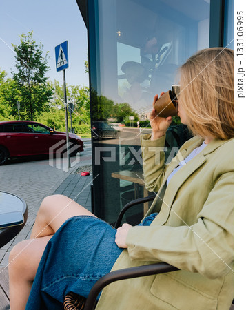 Young blonde businesswoman wearing light green blazer and denim skirt is sitting at outdoor cafe drinking coffee and enjoying moment of peace in urban environment. Woman sipping coffee at outdoor cafe Young blonde businesswoman wearing light green blazer and denim skirt is sitting at outdoor cafe drinking coffee and enjoying moment of peace in urban environment. Woman sipping coffee at outdoor cafe 133106995