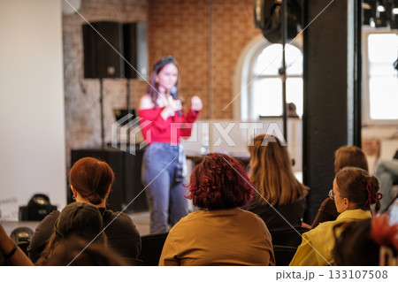 Woman speaks on stage in red off shoulder top to audience during indoor event 133107508