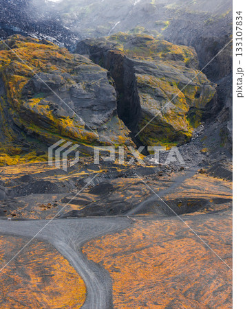 A dramatic Icelandic canyon with steep moss covered cliffs, a gravel path cutting through orange tinged terrain, and dark volcanic rock formations. A dramatic Icelandic canyon with steep moss covered cliffs, a gravel path cutting through orange tinged terrain, and dark volcanic rock formations. 133107834