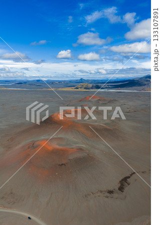 Barren volcanic terrain in Iceland with reddish orange craters, distant mountains, and a small vehicle on a dirt road under a blue sky. Barren volcanic terrain in Iceland with reddish orange craters, distant mountains, and a small vehicle on a dirt road under a blue sky. 133107891