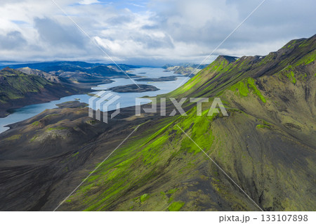 Aerial view of green moss covered hills and dark volcanic terrain in Iceland, with elongated lakes and steep mountain ridges under a partly cloudy sky. 133107898