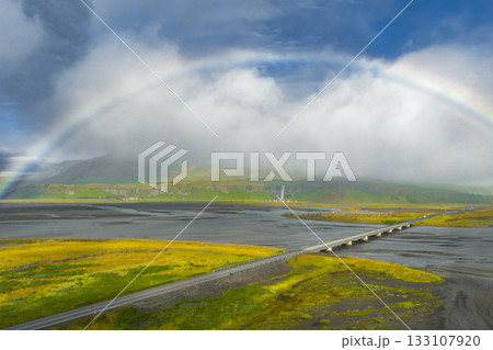 Aerial view of a bridge crossing a wide river in Iceland, surrounded by mossy terrain. A full rainbow arcs above, with clouds over distant cliffs. Aerial view of a bridge crossing a wide river in Iceland, surrounded by mossy terrain. A full rainbow arcs above, with clouds over distant cliffs. 133107920