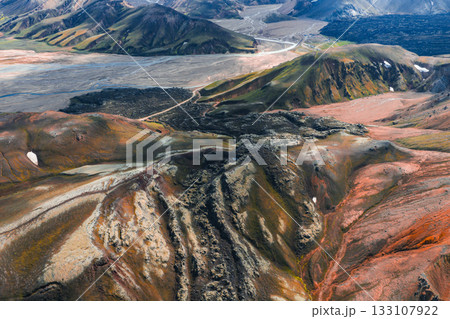 Aerial view of Iceland's Landmannalaugar region with colorful rhyolite mountains, snow patches, volcanic rocks, and a winding dirt road leading to a plain. 133107922
