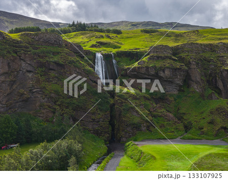 A waterfall flows down a rocky cliff surrounded by green hills under a cloudy sky. A stream winds through the grassy terrain near a visible pathway. 133107925