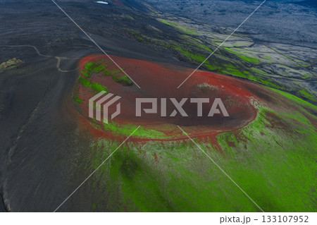 Aerial image of a volcanic crater in Iceland with vivid red and green tones, surrounded by black terrain, a dirt road, and distant rugged landscapes. 133107952