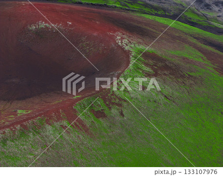 A striking aerial view of a volcanic crater in Iceland, featuring vivid red and brown hues, lush green moss, and rugged surrounding terrain. 133107976