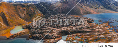 Aerial view of a volcanic terrain in Iceland with dark lava fields, turquoise and green lakes, multicolored hills, and snow capped mountains in the background. 133107981