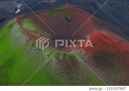 A striking aerial view of a volcanic crater in Iceland, featuring red tones in the interior, green moss on slopes, and a path in the background. 133107987
