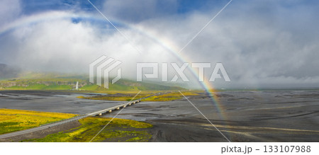 A rainbow arches over black sand and braided rivers in Iceland. A bridge spans the landscape, with a distant waterfall cascading down a misty cliff. A rainbow arches over black sand and braided rivers in Iceland. A bridge spans the landscape, with a distant waterfall cascading down a misty cliff. 133107988