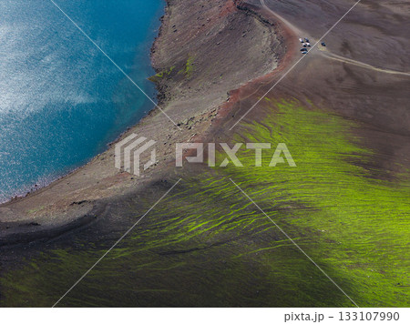 A vivid turquoise lake with a rugged reddish brown shoreline, bordered by green mossy terrain and volcanic soil, seen from an aerial perspective. 133107990