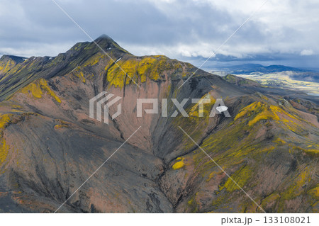 Aerial view of a rugged volcanic mountain in Iceland with yellow green moss, dark rock, a snow patch, rolling hills, and distant plains under cloudy skies. Aerial view of a rugged volcanic mountain in Iceland with yellow green moss, dark rock, a snow patch, rolling hills, and distant plains under cloudy skies. 133108021