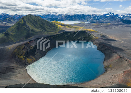 Aerial view of a vibrant blue crater lake in Iceland, surrounded by dark volcanic terrain, a green mountain, snow capped peaks, and a partly cloudy sky. 133108023